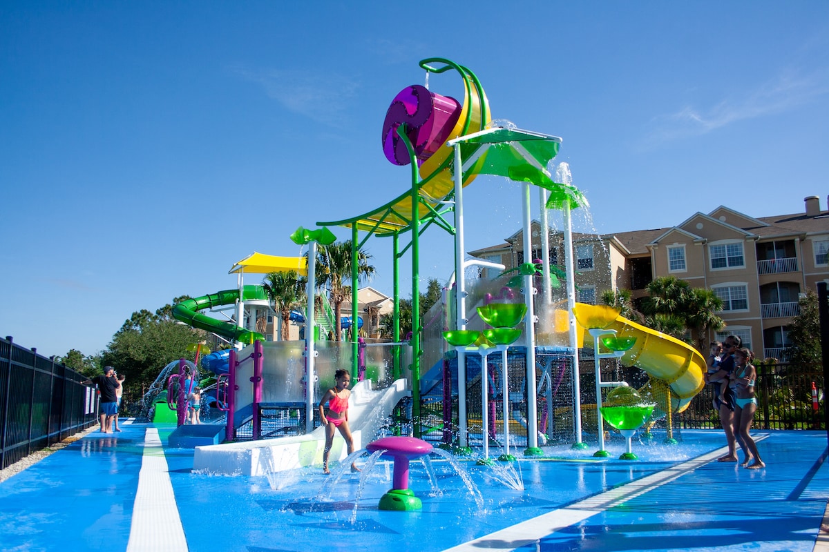 A vibrant water play area features colorful slides and fountains, offering a fun and engaging experience for children. The blue surface reflects sunlight, enhancing the cheerful ambiance. Palm trees and resort buildings are visible in the background, contributing to the leisure environment.