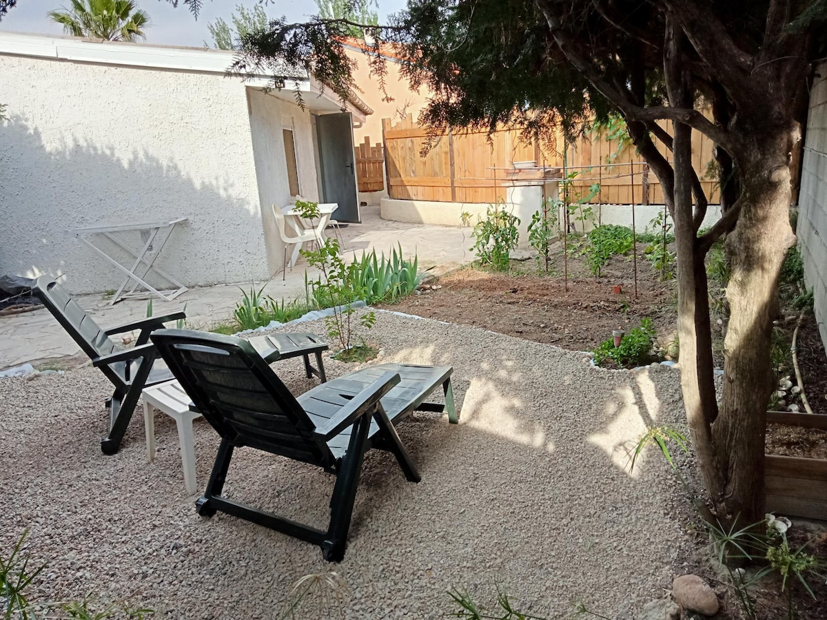 An outdoor area features two lounging chairs positioned on gravel, surrounded by a tranquil garden. Green plants and small flowers are visible, with a wooden fence providing privacy. In the background, a small building is partially visible under clear skies.