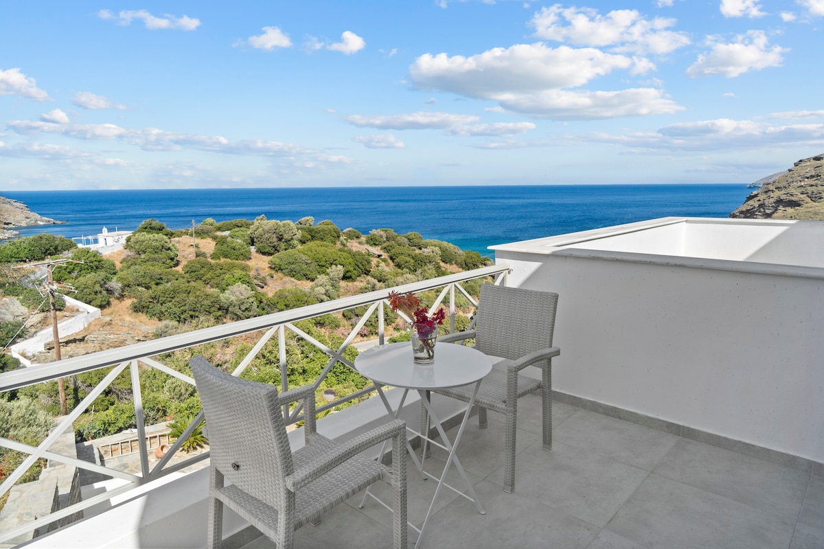 A balcony offers a scenic view of the Aegean Sea and surrounding greenery. Two wicker chairs are positioned around a small round table, creating a cozy space for relaxation. The clear blue sky complements the tranquil atmosphere of the outdoor setting.