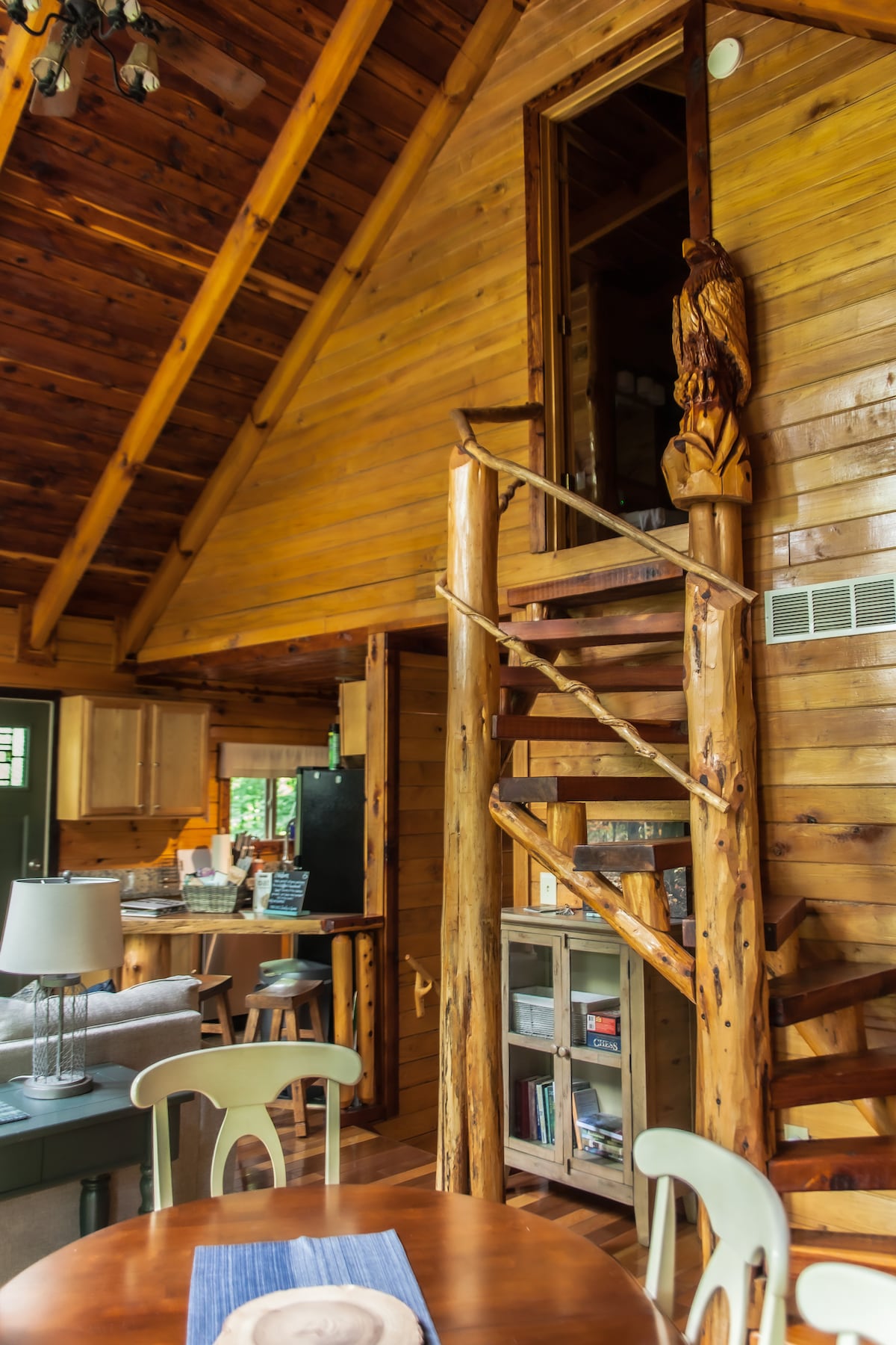 A wooden spiral staircase is seen, leading to a loft area adorned with natural wood finishes. The surrounding cabin features exposed wood beams and a dining table set nearby. A glimpse of the kitchen can be seen in the background, providing an inviting ambiance.
