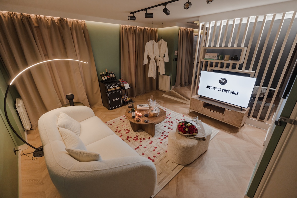 A modern living area is presented with a light-colored sectional sofa and a wooden coffee table adorned with decorative candles and flowers. Soft curtains frame the space, while a large television is positioned in front of a stylish partition, providing a cozy atmosphere.