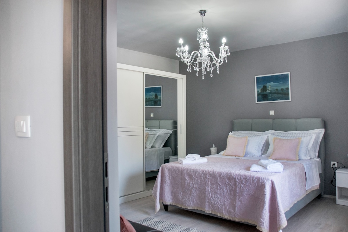 A spacious bedroom is illuminated by a chandelier. The bed is dressed in light-colored linens, with pillows arranged at the head. A mirrored closet reflects the room, complemented by a framed artwork on the wall. Soft natural light filters through the space.