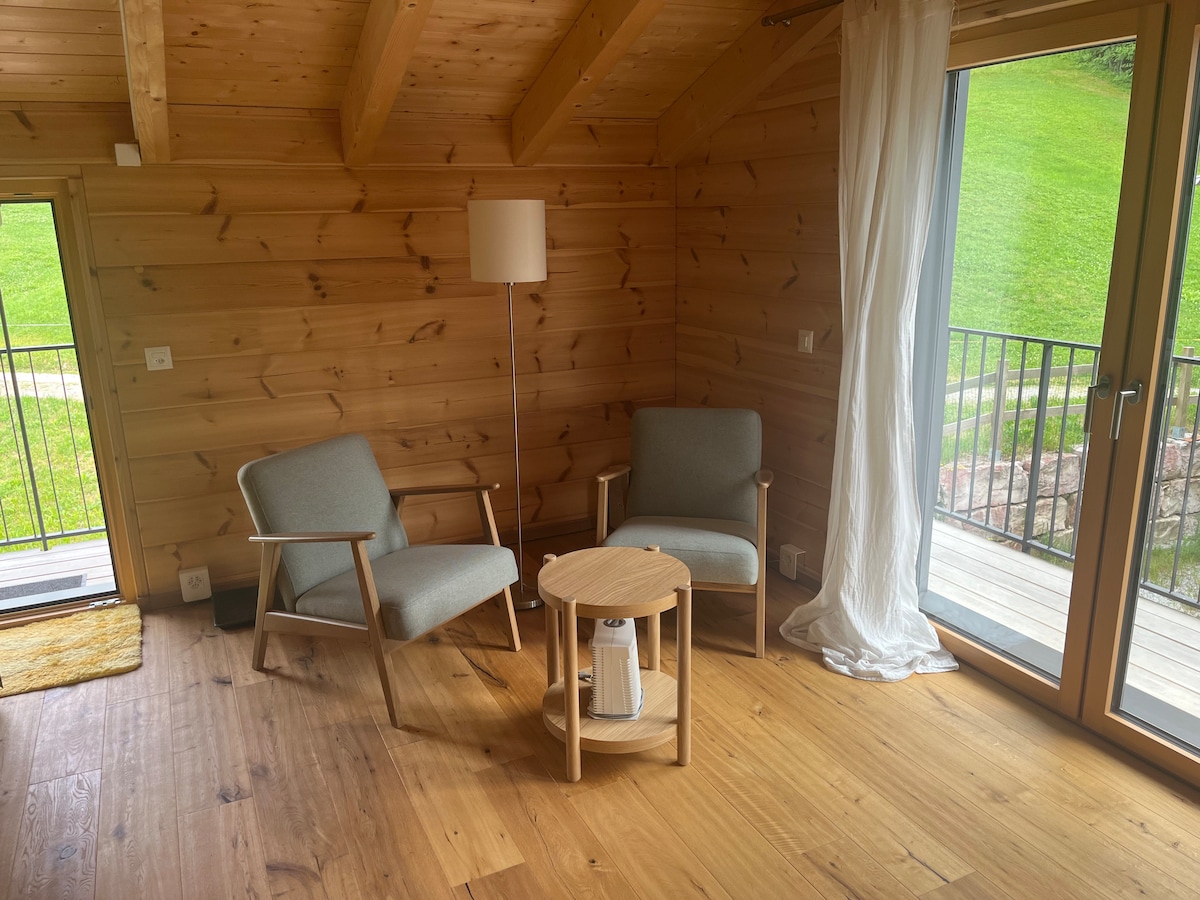 A cozy seating area is featured, comprising two light gray chairs with wooden frames and a small round wooden table. Natural light enters through large glass doors, which open to a grassy exterior. The wooden walls and floor create a warm ambiance.