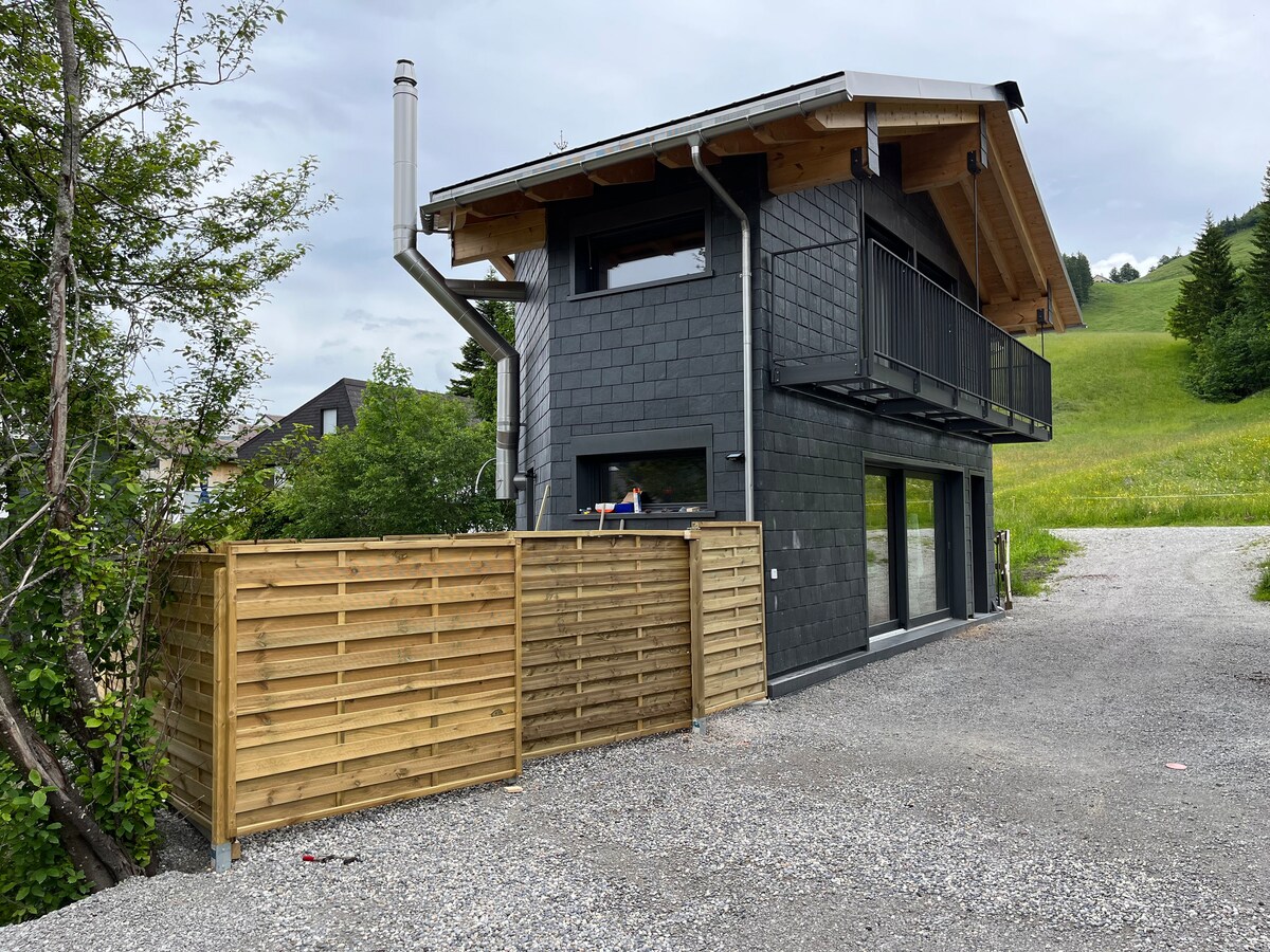 A modern structure is showcased, featuring dark exterior siding and a spacious balcony. The two-story building is set against a green hillside, with a gravel pathway leading to its entrance. Surrounding foliage and wooden fencing enhance the natural landscape.