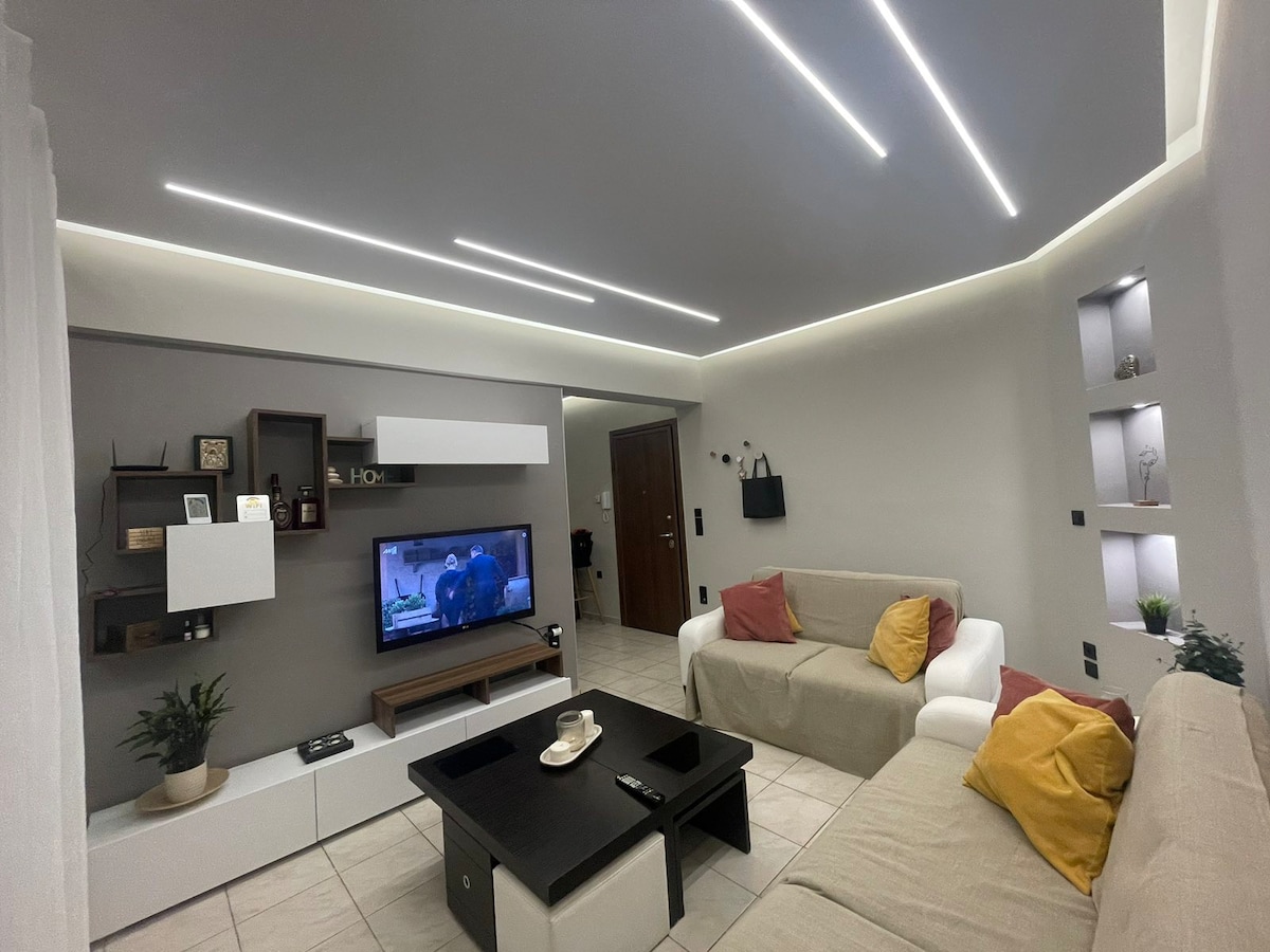 A modern living room is presented, featuring a light-colored sofa and armchair accented with colorful cushions. A wall-mounted TV is positioned against a neutral wall, complemented by decorative shelving. Soft lighting enhances the room's contemporary design, with a coffee table at the center.