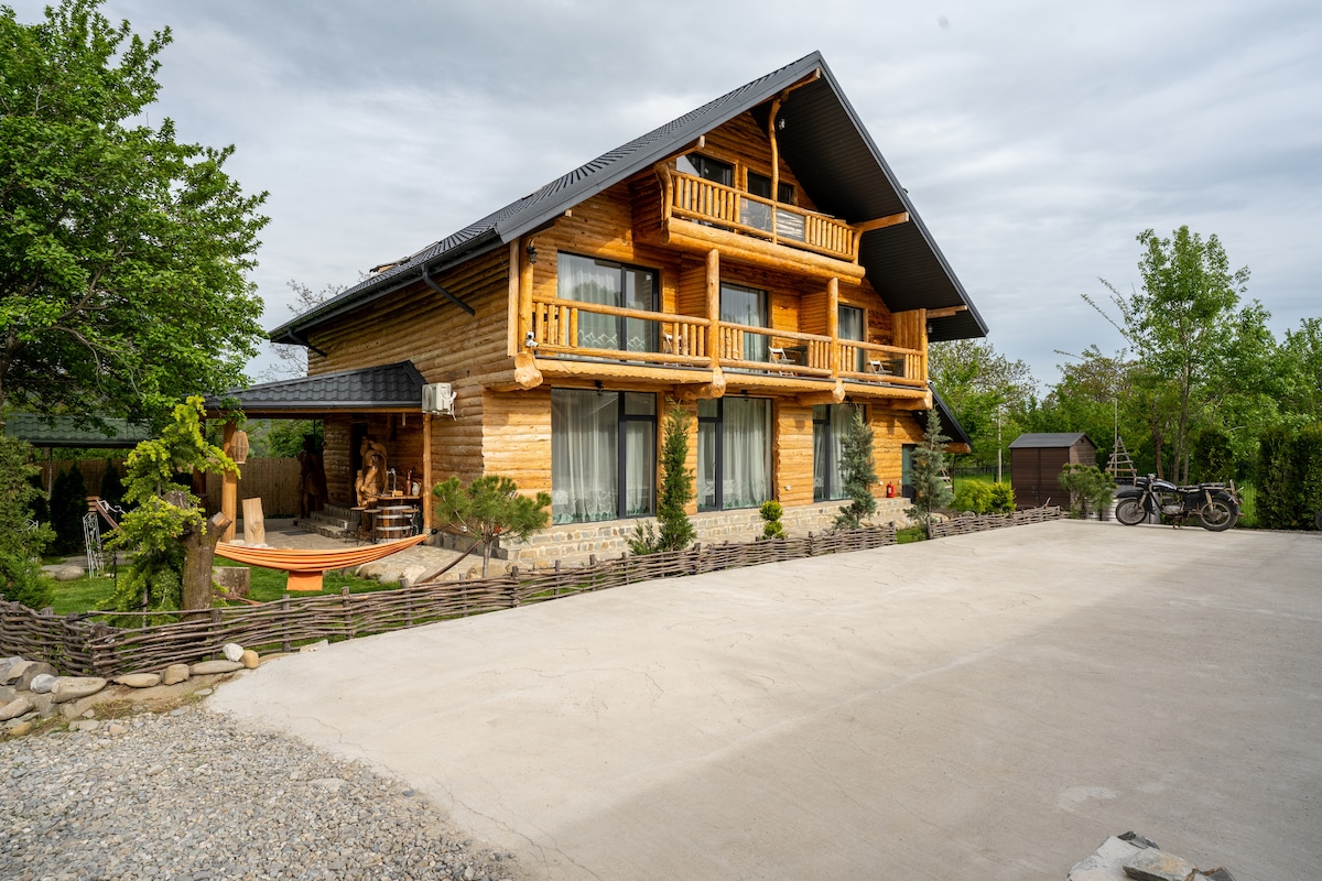 A spacious wooden house is showcased, featuring multiple balconies with wooden railings. The landscape includes greenery and a gravel area, while a parking space is visible in the foreground. Decorative elements, such as rustic sculptures, enhance the outdoor environment.