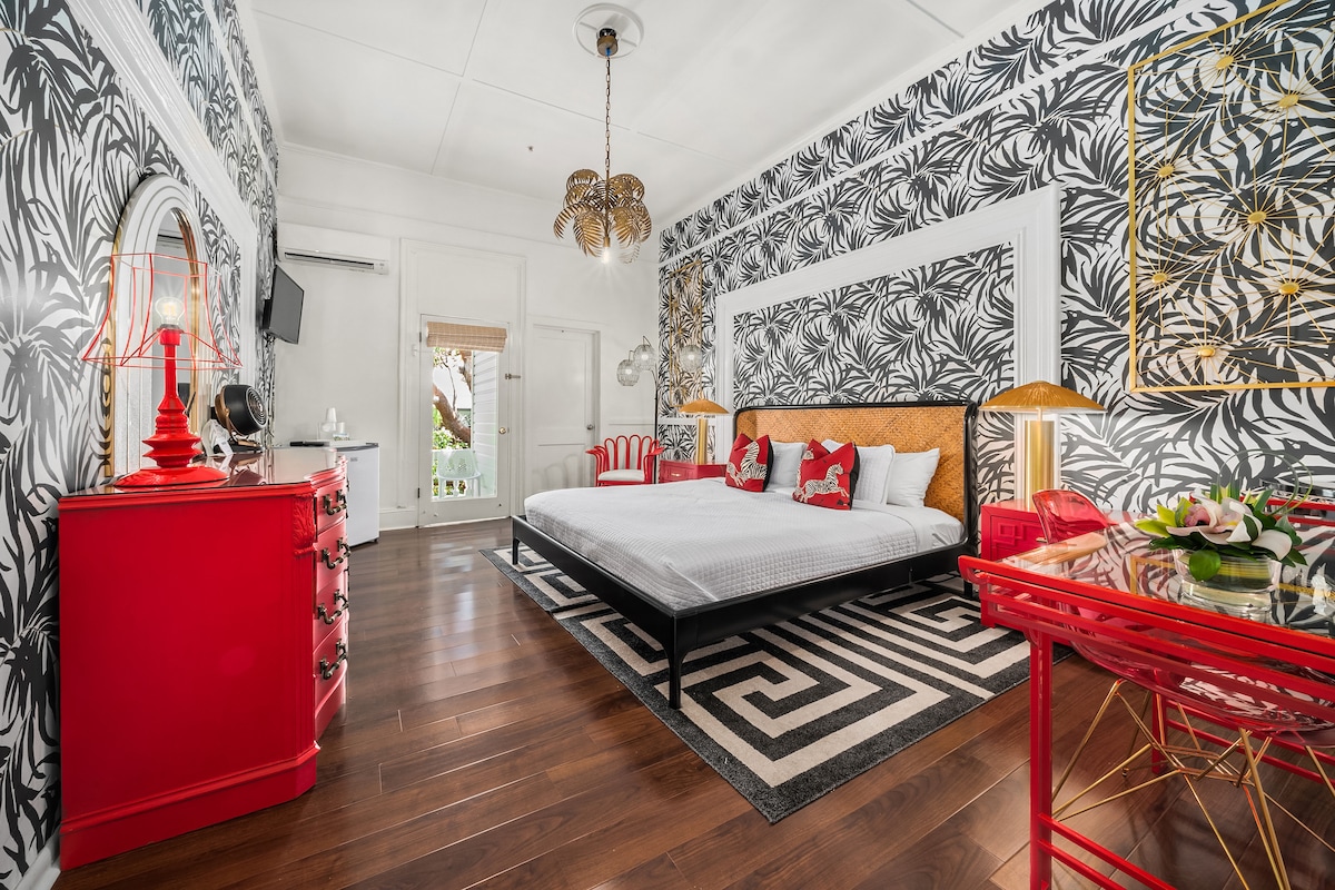 A stylish room features a king bed with crisp linens centered against a bold black and white patterned wall. A chic red dresser and a glass desk complement the decor. Natural light filters through the window, enhancing the room's vibrant atmosphere.