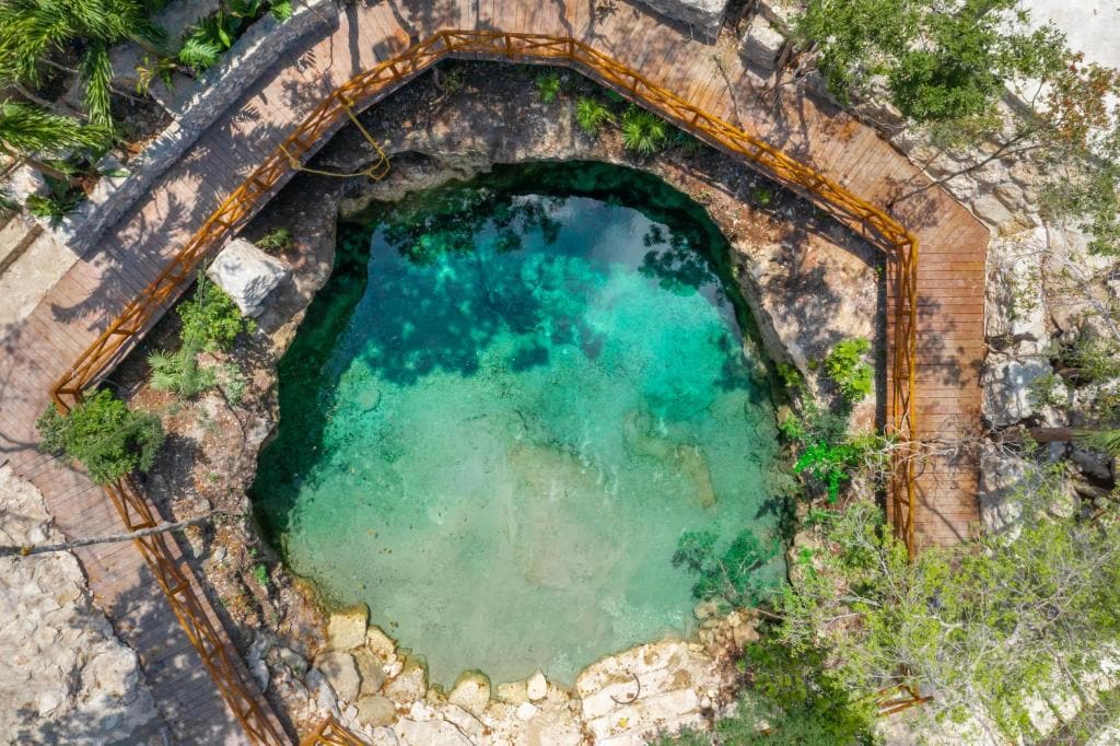 An aerial view captures a natural cenote surrounded by a wooden deck and lush greenery. The clear, turquoise water reveals a rocky bottom, while the surrounding trees provide shade, creating a serene environment for relaxation and enjoyment.