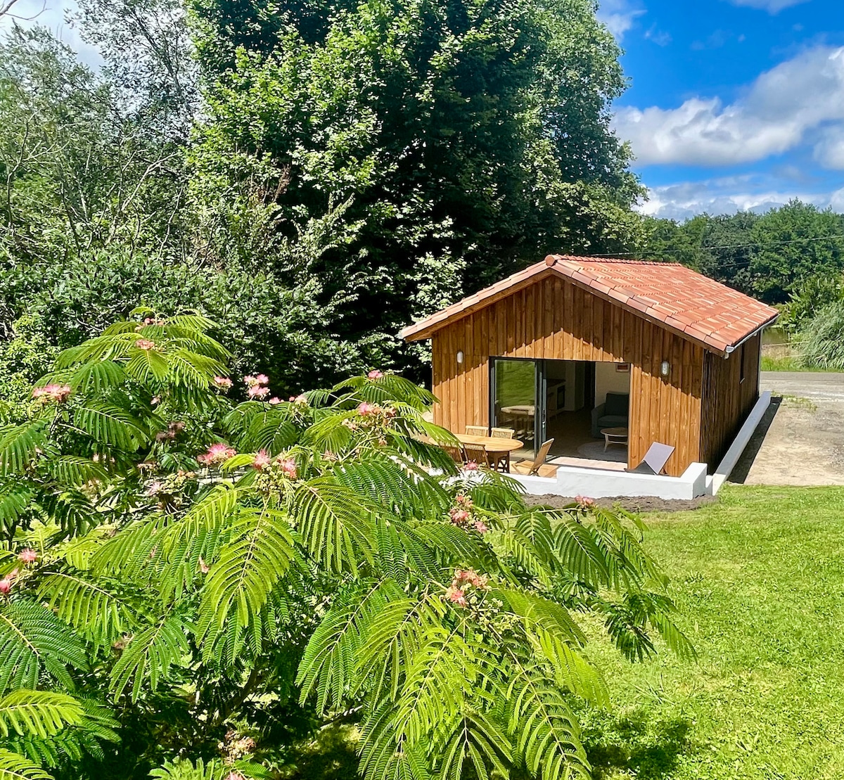 A wooden lodge is surrounded by lush greenery, featuring a sloped roof and expansive glass doors that invite natural light. Outdoor seating is visible on the terrace, with a comfortable chair positioned near the entrance, offering views of the serene landscape.