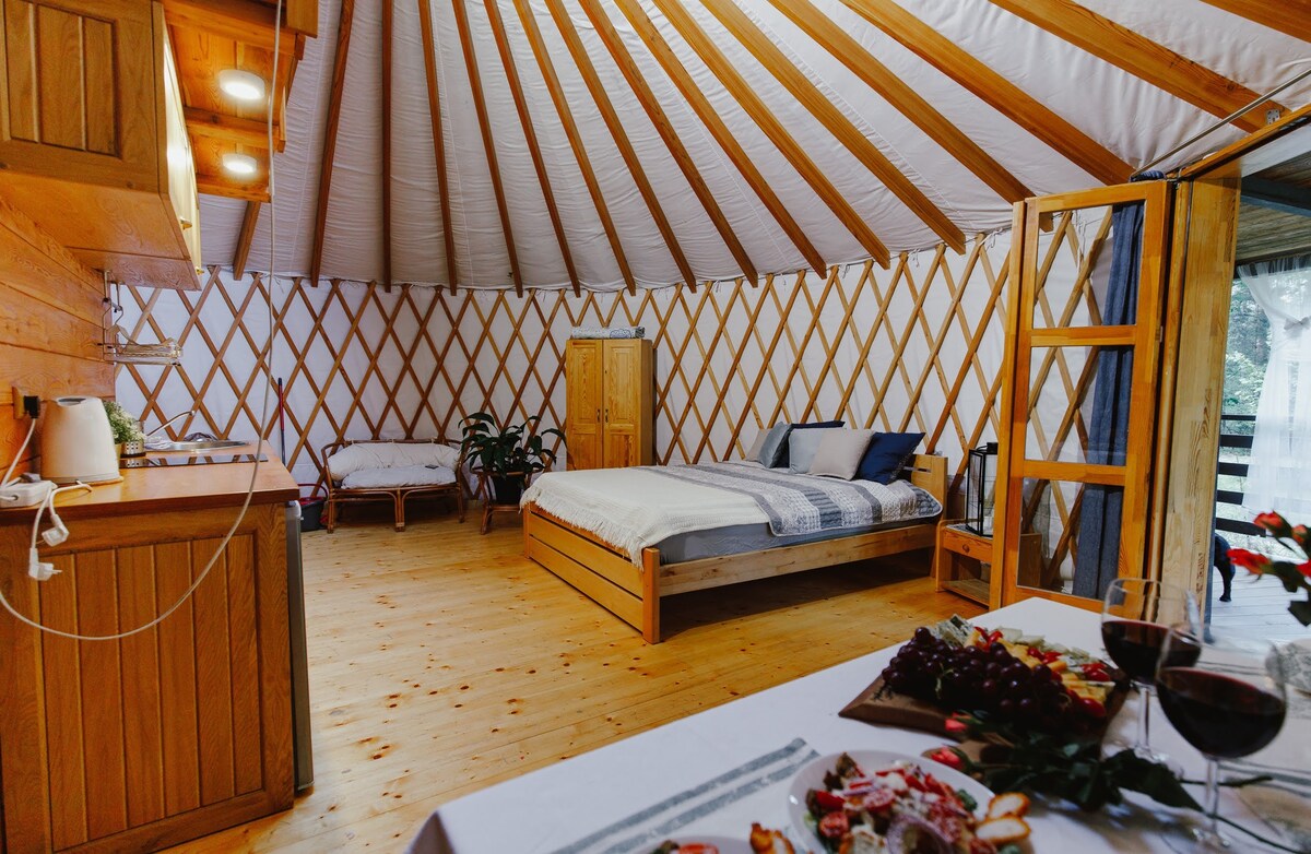 A spacious interior features a wooden yurt with a large bed and a cozy seating area. Natural wood accents are highlighted by soft lighting. A small kitchenette is visible, and a dining table is set with snacks and drinks, providing a welcoming atmosphere.