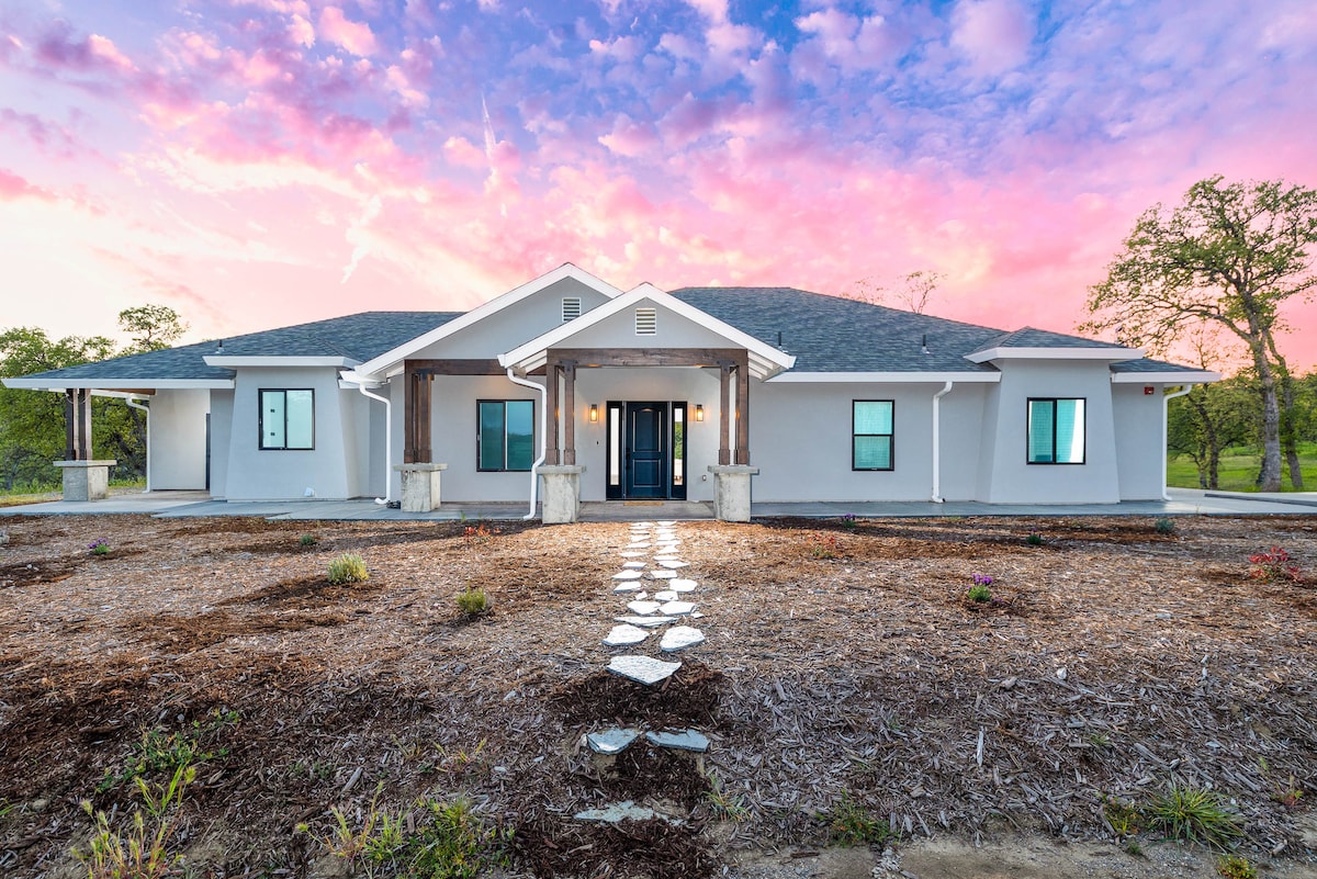 A modern home is set against a colorful sunset, featuring a welcoming entrance with a covered porch. The pathway of stepping stones leads to the central door, flanked by large windows that provide a view of the landscaped yard.