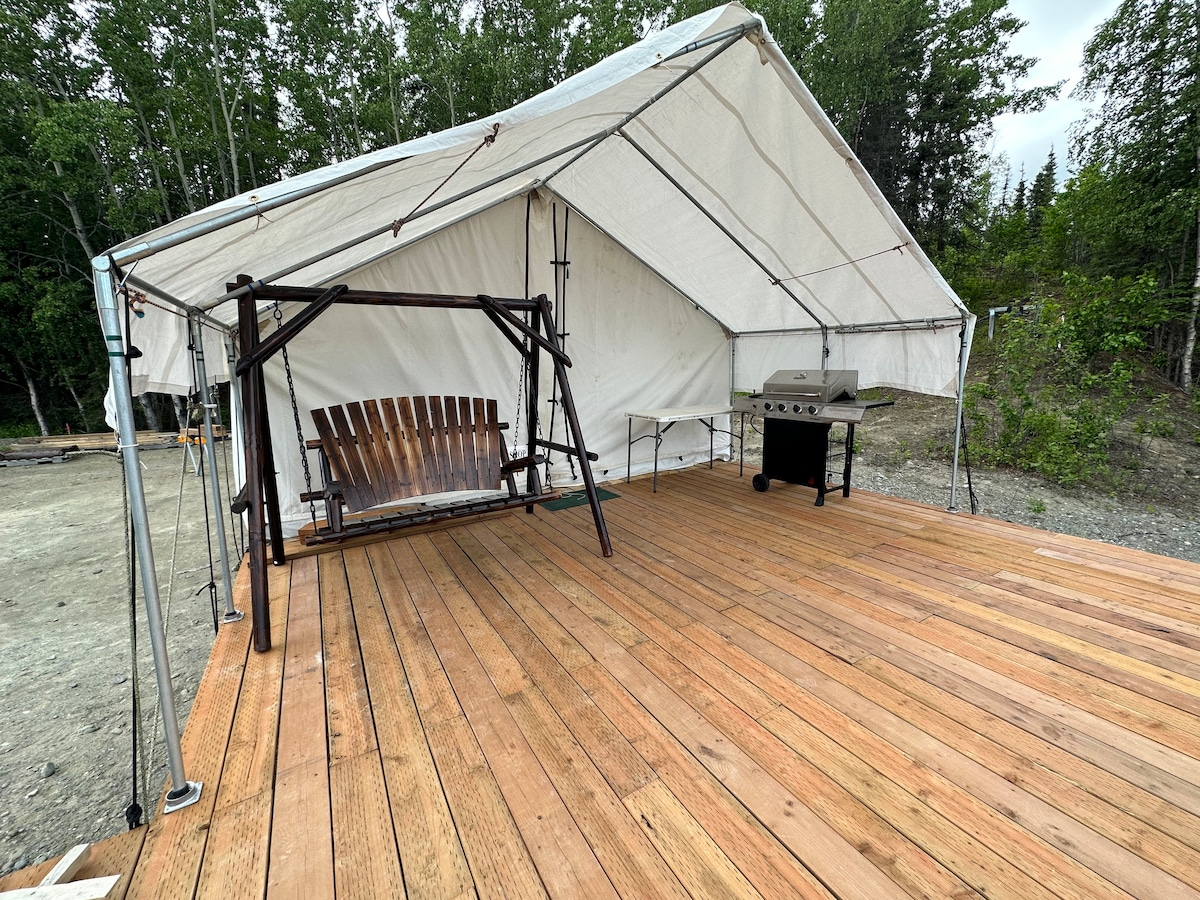 A spacious wooden deck is presented under a large white tent, featuring a wooden swing and a barbecue grill. Surrounding greenery is visible, creating a natural setting that enhances the outdoor living area.