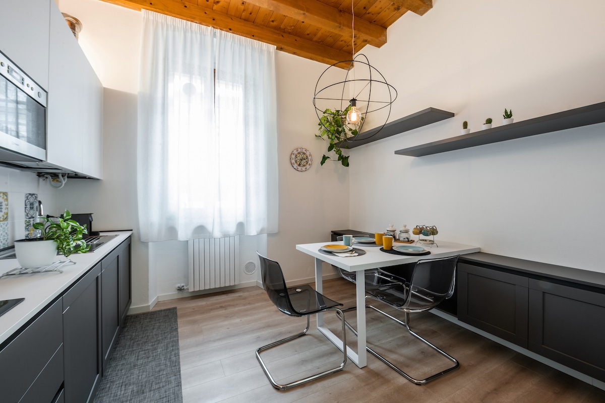 The kitchen area features modern cabinetry in neutral tones, complemented by a white table surrounded by sleek black chairs. A large window allows natural light to brighten the space, which is adorned with green plants and minimalist decor. Light wooden ceilings add warmth to the atmosphere.