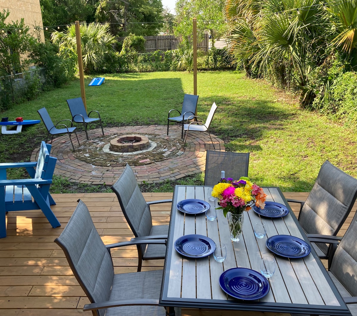 An outdoor dining area features a table set for six, complete with plates and glasses. Surrounding a circular fire pit built with bricks, four chairs are positioned, while a green lawn and palm trees create a tranquil atmosphere.