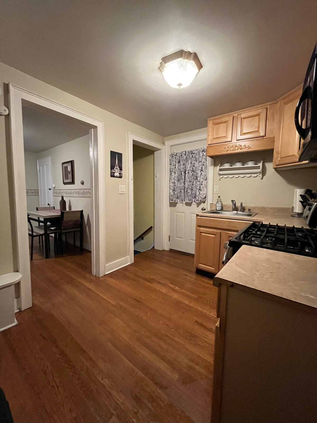 The kitchen is designed with wooden cabinetry and a spacious countertop, featuring a gas stove and overhead microwave. A doorway leads to a dining area, while light is provided by a ceiling fixture and window treatments. Warm wooden flooring complements the neutral wall colors.