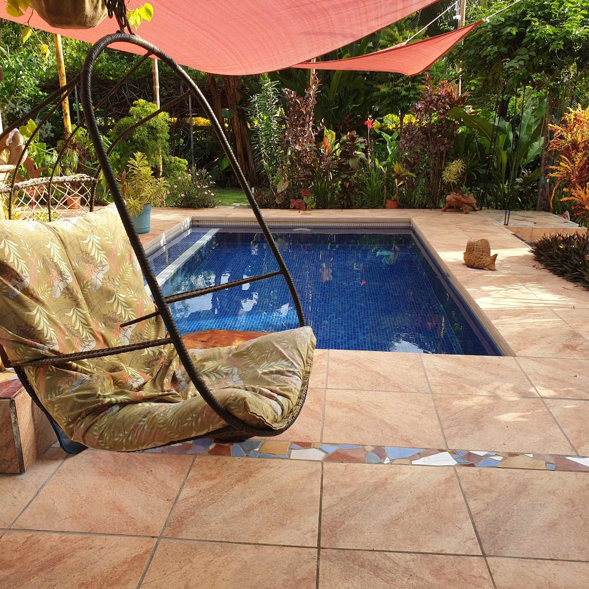 A swinging chair with a leafy pattern is positioned beside a tranquil pool, which features clear blue water. The outdoor area is surrounded by lush greenery and colorful plants, complemented by a terracotta tiled surface. Shade is provided by decorative fabric overhead.