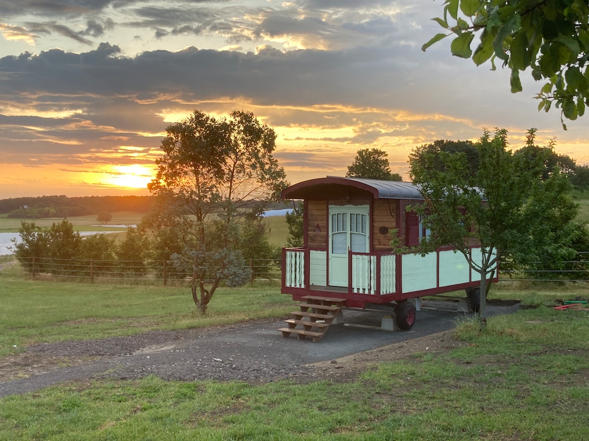 A charming roulotte is set against a backdrop of a vibrant sunset, with soft hues illuminating the sky. The structure is surrounded by green grass and trees, offering a tranquil outdoor setting. Stairs lead up to the entrance, framed by a welcoming porch.