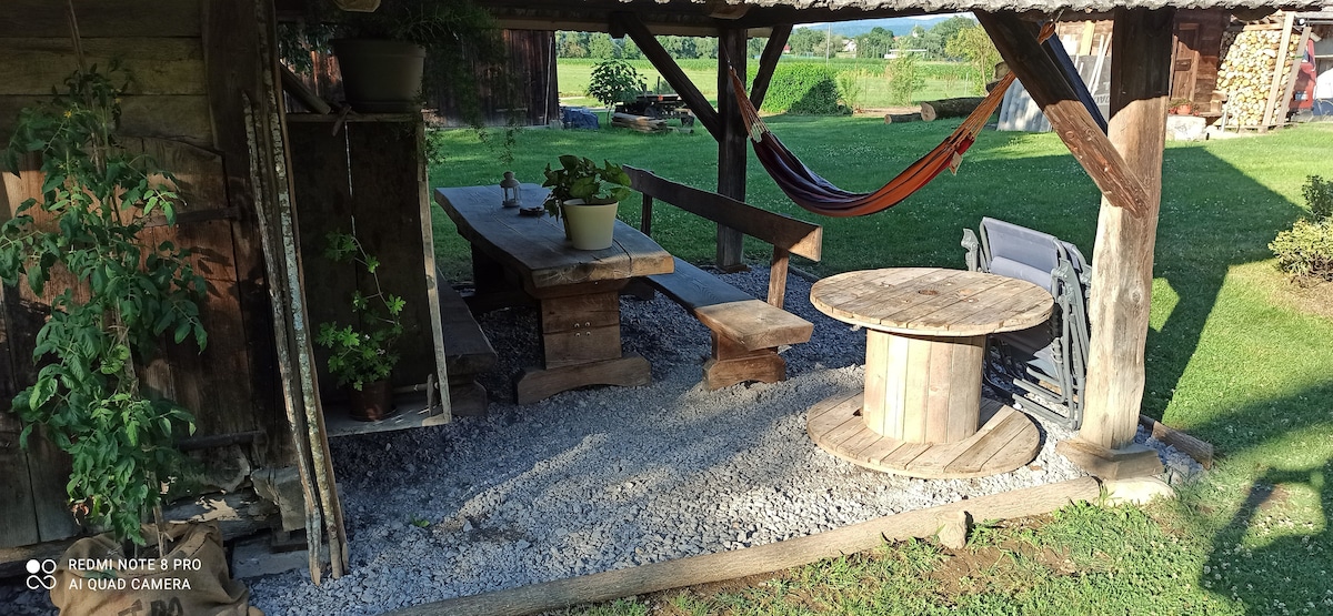 An outdoor seating area is presented, featuring a wooden table and benches surrounded by lush greenery. A hammock is seen hanging nearby, offering a relaxed spot for rest. Decorative plants are placed on the table, and gravel covers the ground, providing a rustic charm.