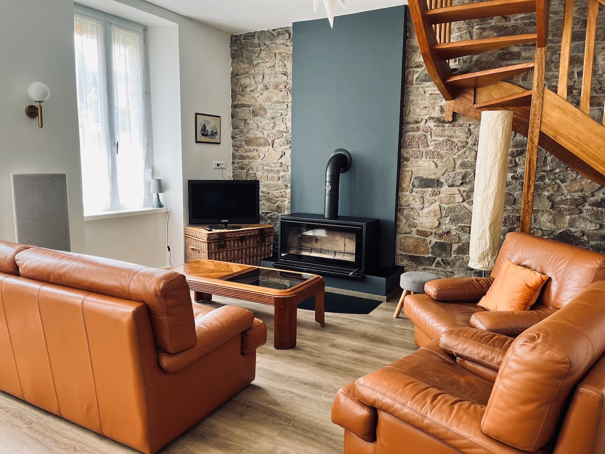 A welcoming living area is highlighted by stone walls and natural light. Two brown leather sofas and a wooden coffee table are arranged around a wood-burning stove, creating a cozy gathering space. A spiral staircase leads to the upper level, enhancing the open layout.