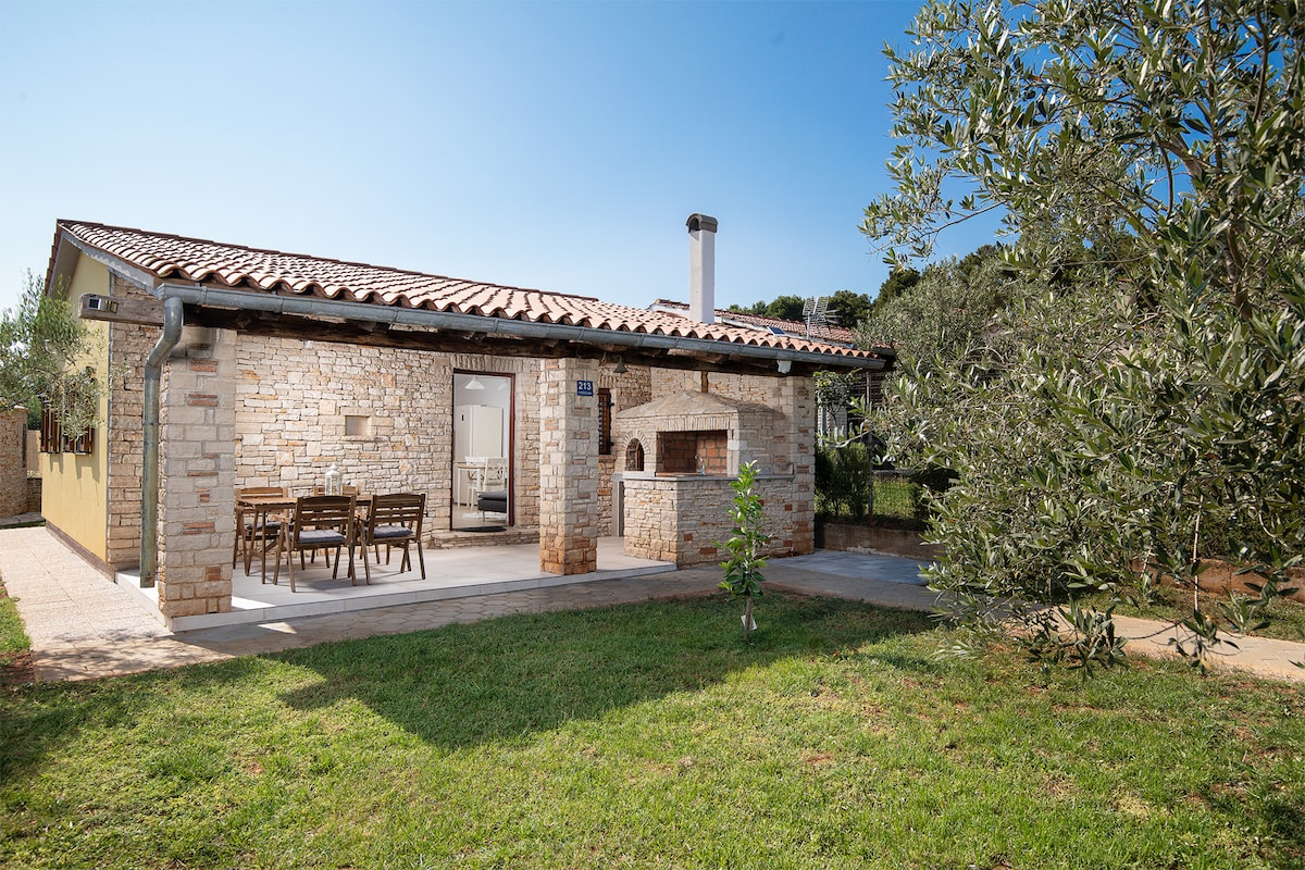 A charming stone house is presented, featuring a covered terrace with a dining area and a built-in outdoor barbecue. Surrounding greenery enhances the tranquil atmosphere, and a well-maintained lawn offers space for relaxation. The structure is complemented by a clay-tiled roof and large glass doors.