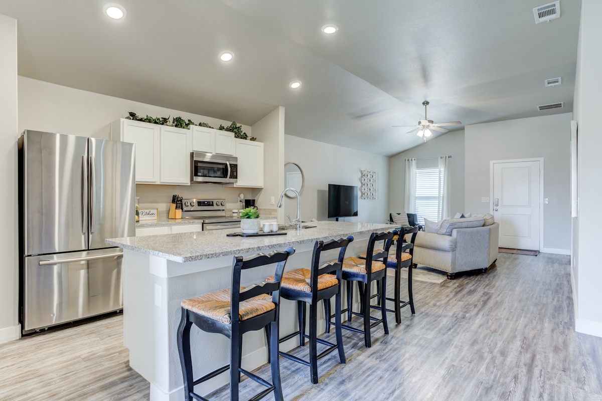 A modern kitchen is showcased, featuring stainless steel appliances and a large island with seating for six. Open to the living area, the space is brightened by recessed lighting and a ceiling fan, while light wooden flooring adds warmth.