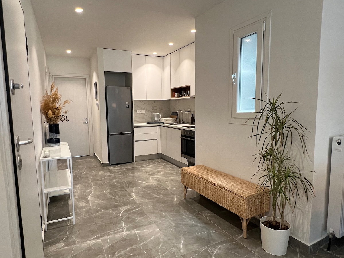 A modern kitchen area features sleek cabinetry and stainless steel appliances, including a refrigerator and oven. The open space is adorned with a light-colored table and a decorative potted plant, complementing the marble-patterned flooring and bright wall finishes.