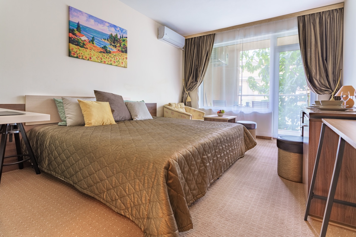 A spacious sleeping area features a large, quilted bed adorned with several decorative pillows. Natural light filters through a large window, illuminating the room. A painting of a landscape is displayed above the bed, and a small sitting area is arranged beside it.