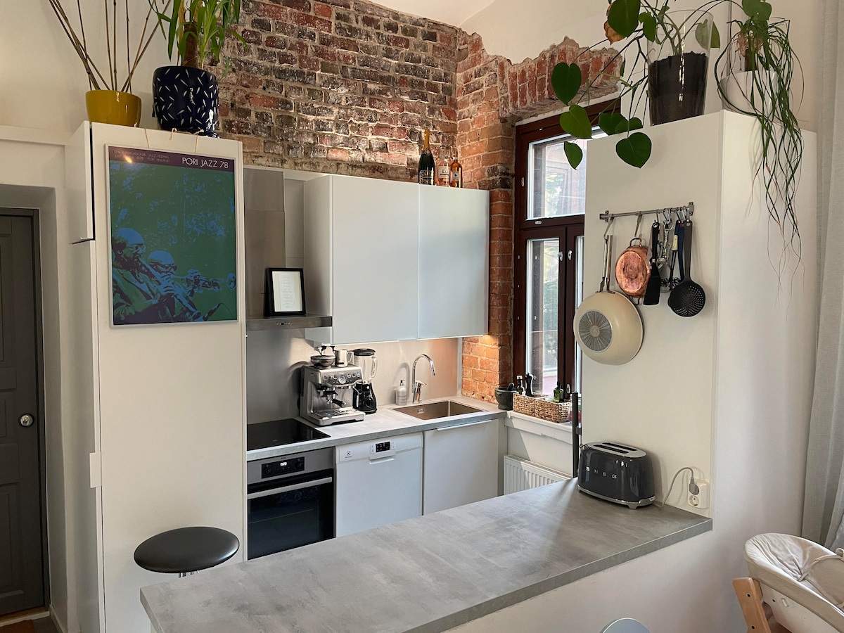 A modern kitchen is featured with sleek white cabinetry and a rustic brick wall, providing a warm contrast. Appliances are integrated, and a compact countertop offers casual seating. Potted plants add a touch of greenery, while artwork decorates the wall, enhancing the space's character.