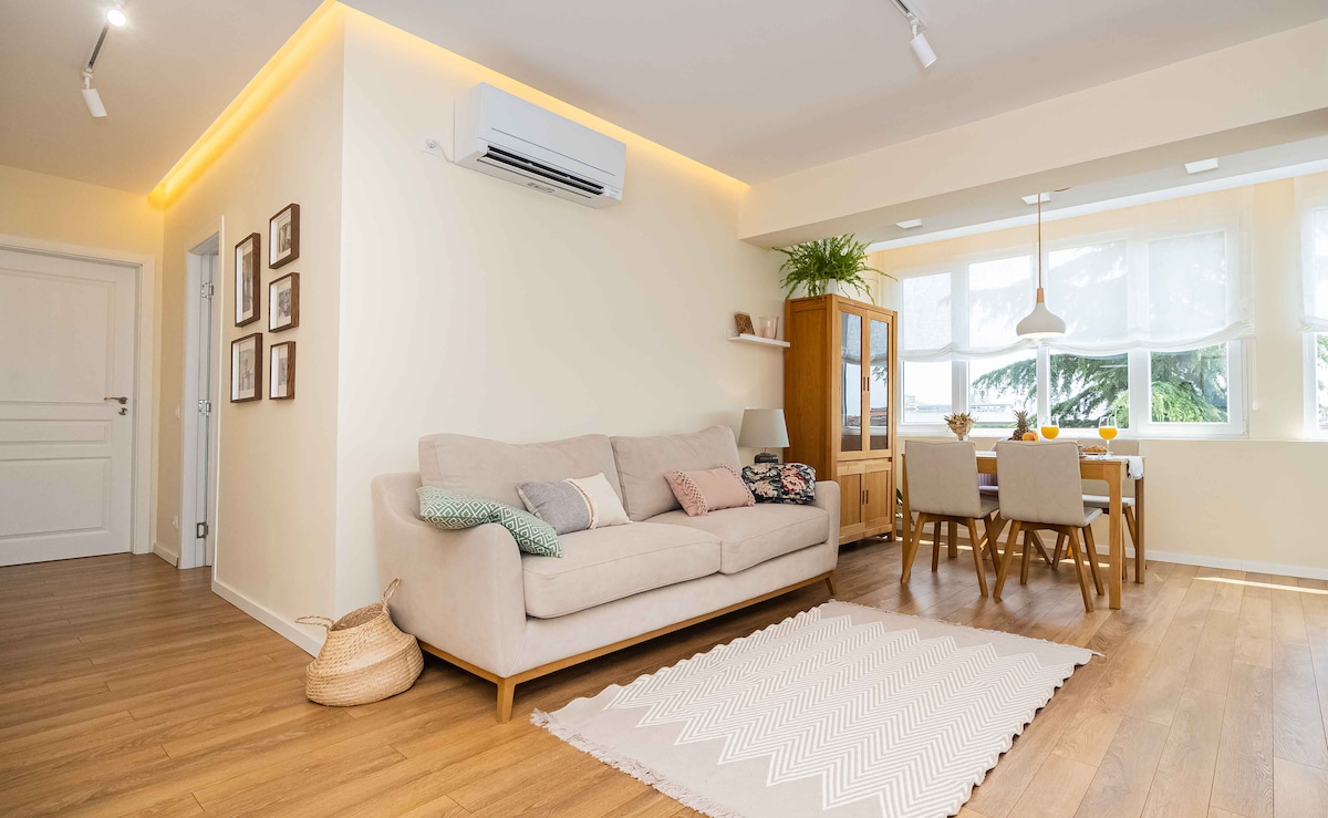A light and airy living space features a soft beige sofa complemented by patterned cushions. Natural wood flooring extends throughout the area, enhanced by warm lighting. A dining table is positioned beside large windows, creating an inviting environment. Decorative plants add a touch of greenery.