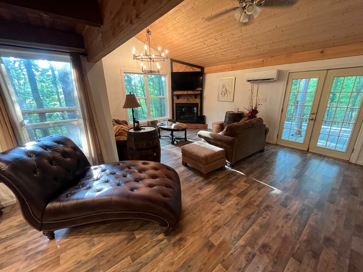 A spacious living area is shown with a leather chaise lounge, a coffee table, and a comfortable couch. Large windows allow natural light to brighten the space, revealing views of the surrounding trees. A decorative chandelier hangs from the ceiling above.