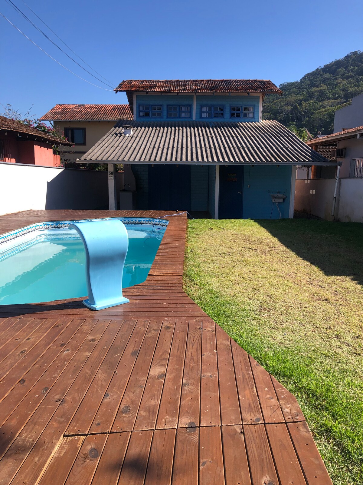A wooden deck surrounds a clear swimming pool set in a green lawn. A two-story house with blue windows and a sloped roof is positioned in the background, complemented by a sunny sky. The area appears spacious and well-lit.
