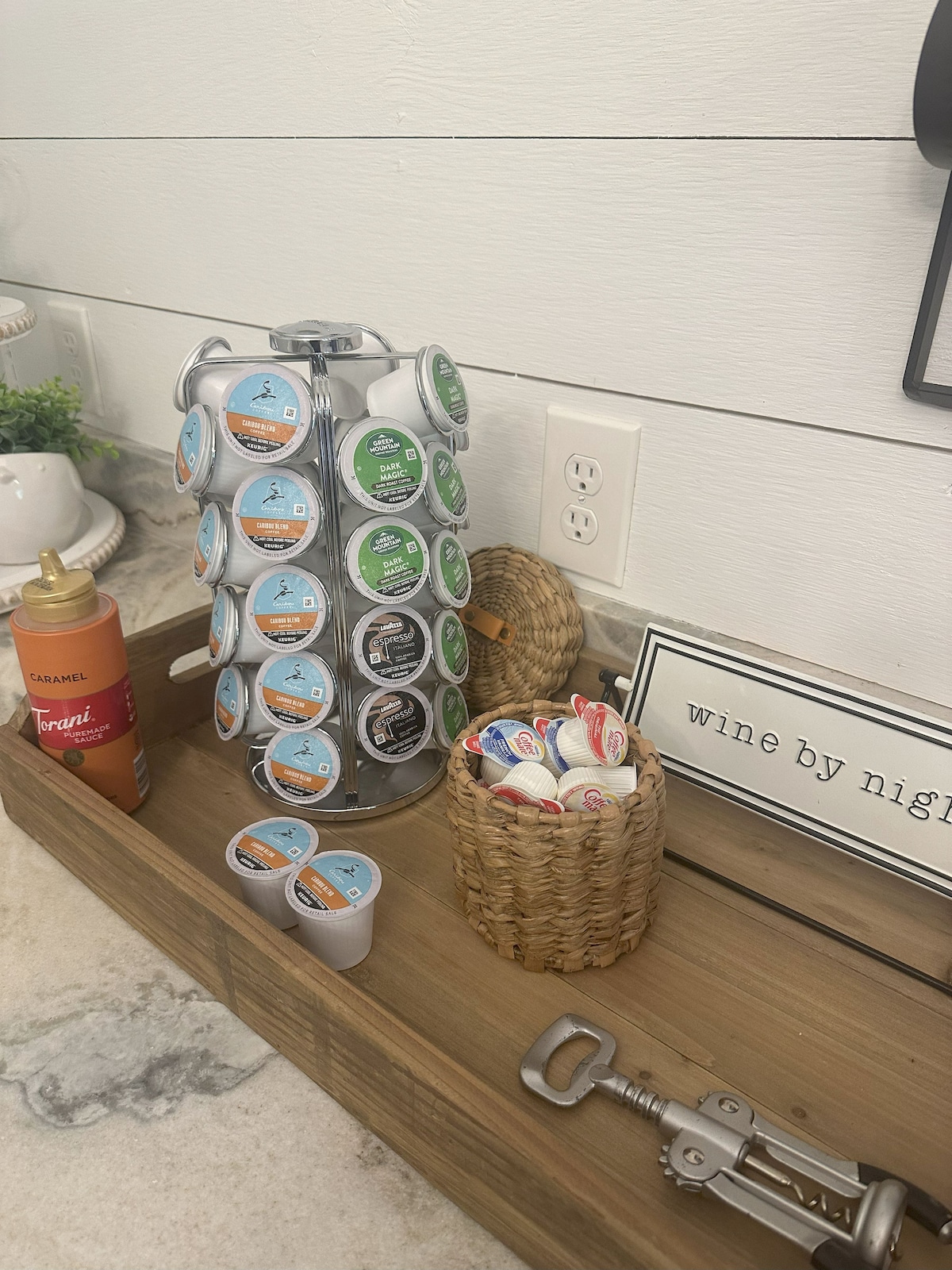 A coffee pod holder showcases a variety of coffee flavors, arranged in a circular display. Nearby, a woven basket contains creamers, while a bottle of syrup adds color. The rustic countertop features wood accents, contributing to the functional kitchen setup.