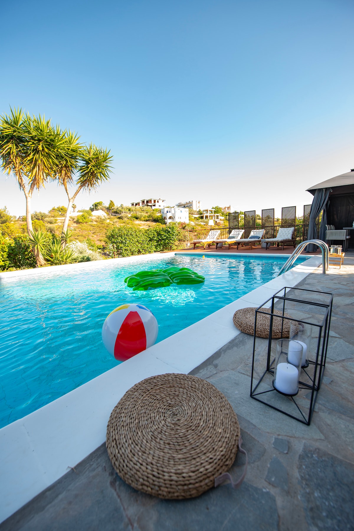 A crystal-clear pool is visible, surrounded by leafy palm trees. Comfortable loungers line the pool's edge. A colorful beach ball floats on the water's surface, adding a playful touch. Natural stone paving and decorative lanterns enhance the outdoor leisure area.