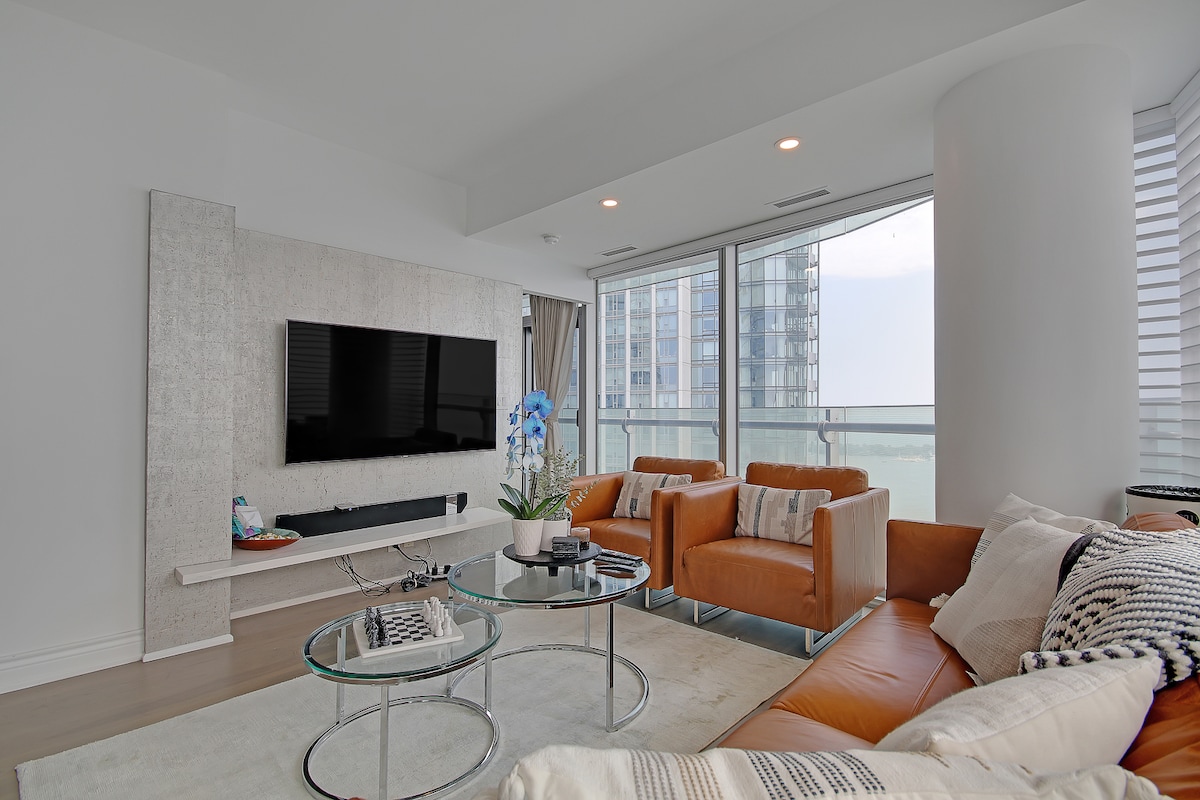 A modern living area features a large wall-mounted television and stylish orange armchairs. A low glass coffee table is surrounded by a light-colored area rug. Floor-to-ceiling windows provide a view of the outside, allowing natural light to fill the space.