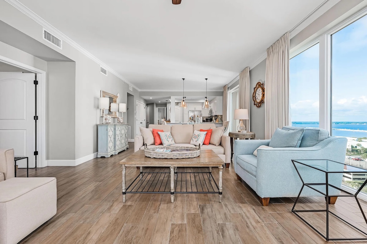 A spacious living area features large windows offering expansive views of the water. Comfortable seating arrangements, including a light blue chair and neutral-toned sofas with decorative pillows, surround a rustic coffee table. Warm wood flooring provides a cozy backdrop to the soft color palette.