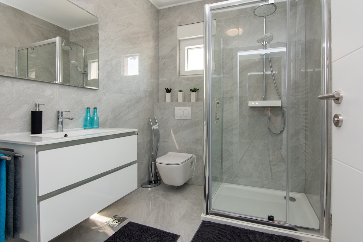 A modern bathroom features sleek grey tiles and a glass shower enclosure. A white vanity with minimalistic design and a large mirror is complemented by blue glass decor. Natural light enters through two small windows, creating a serene and functional space.
