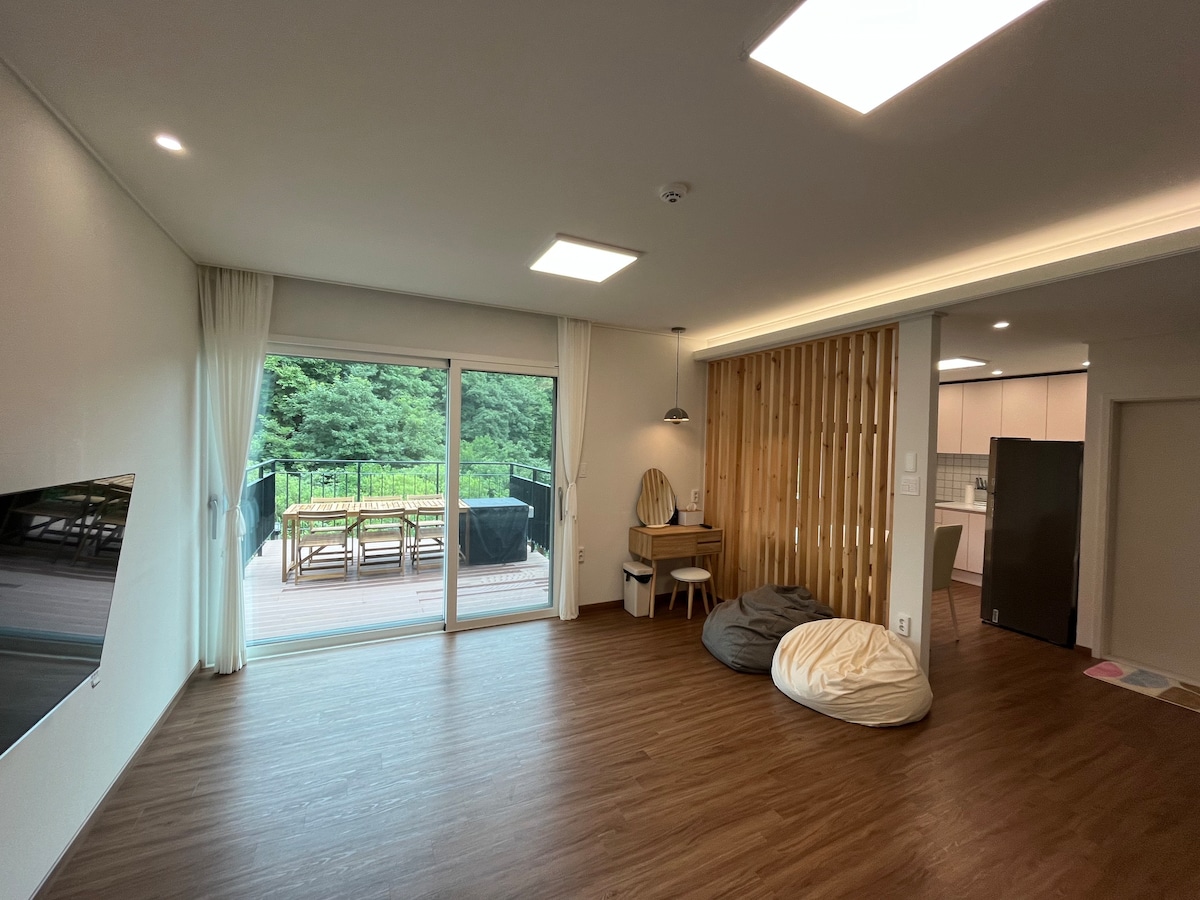 A spacious living area features large sliding glass doors that open to a private deck surrounded by trees. Light-colored walls and wood flooring create a warm ambiance. A dining table and chairs are positioned along with two cozy bean bags, offering additional seating options.