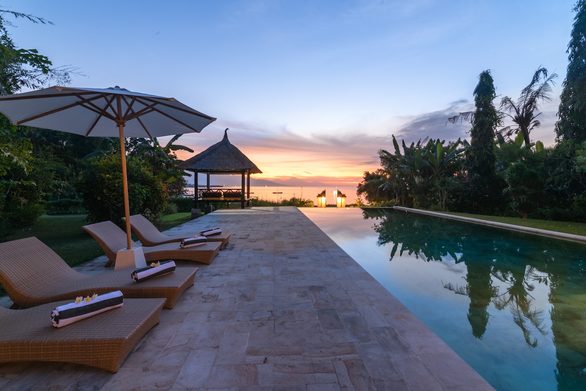An infinity pool reflects the colors of a sunset, bordered by a spacious stone deck. Four lounge chairs are positioned nearby, and a pavilion with a thatched roof is visible in the background, surrounded by lush greenery and tropical plants.