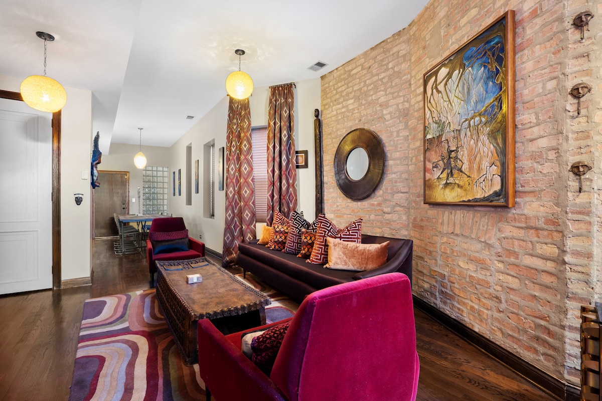 The living area features a blend of warm tones and unique textures. A dark sectional sofa adorned with colorful cushions is positioned across from a wooden coffee table. A vibrant area rug adds depth to the space, while a piece of wall art provides a focal point.
