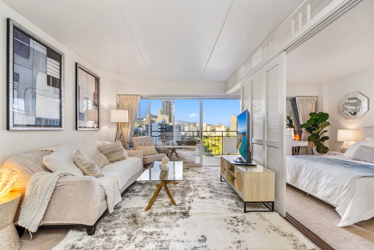 The living area features a comfortable light-colored sofa, accompanied by a glass coffee table atop a textured rug. A large swivel TV is positioned across from the seating. Sliding doors lead to a bright bedroom space, which includes a spacious bed, adorned with soft linens.