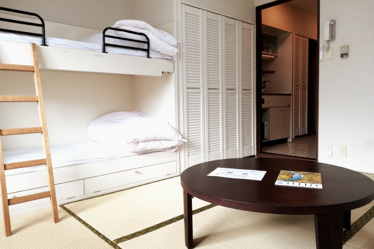 A room features a cozy bunk bed with white bedding and a wooden ladder. A low wooden table sits on tatami flooring, providing a traditional touch. Closet doors line the wall, leading towards a doorway that offers glimpses of a kitchenette in the background.