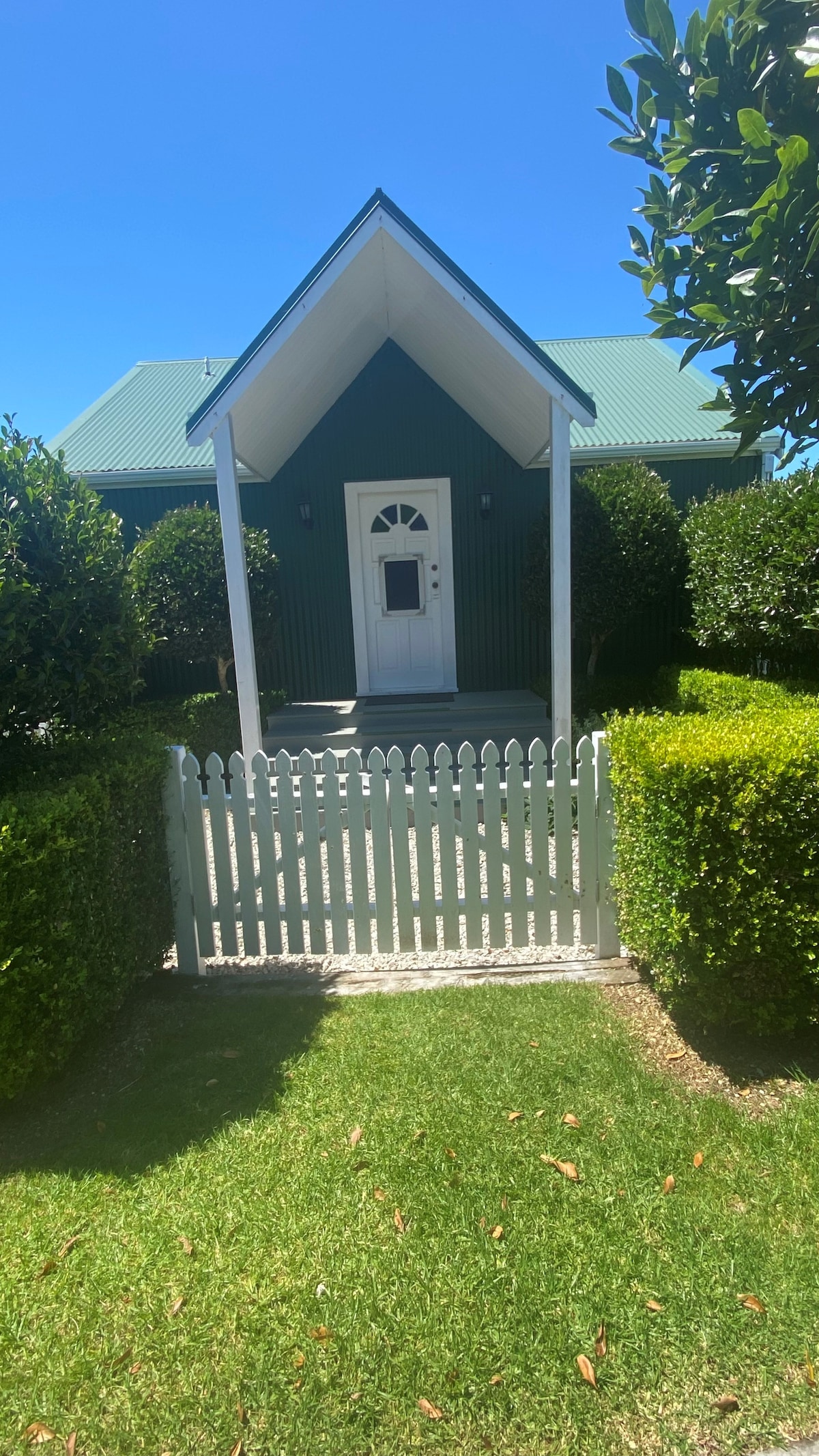The entrance features a quaint white picket fence leading to a green cottage with a pointed roof. Neatly trimmed hedges flank the path, and sunlight illuminates the well-maintained lawn, creating a welcoming approach to the home.