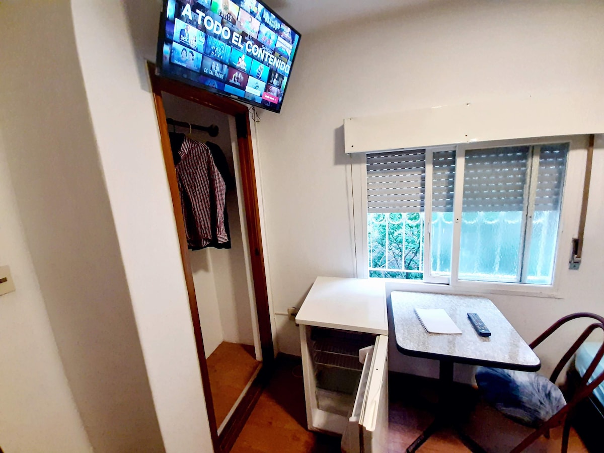 A corner of the room features a smart TV mounted on the wall, providing entertainment options. A small table is positioned beside a chair, suitable for work or dining. Natural light enters through the window, which is covered by blinds, enhancing the gentle ambiance.