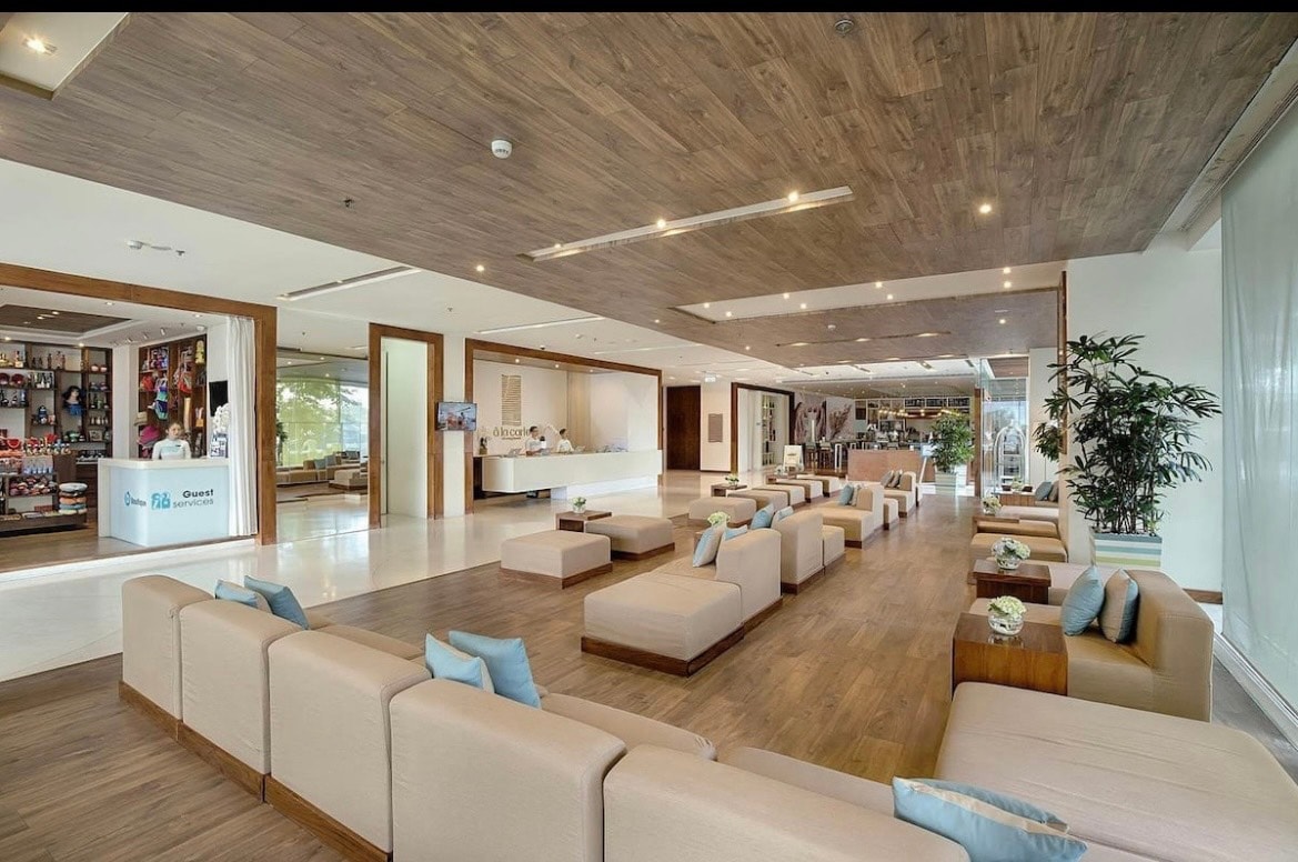 A spacious lobby area features light-colored seating arrangements with natural wood accents. Soft, neutral tones are complemented by greenery and decorative elements. Natural light floods through large windows, creating a bright and welcoming environment. A reception desk is visible in the background.