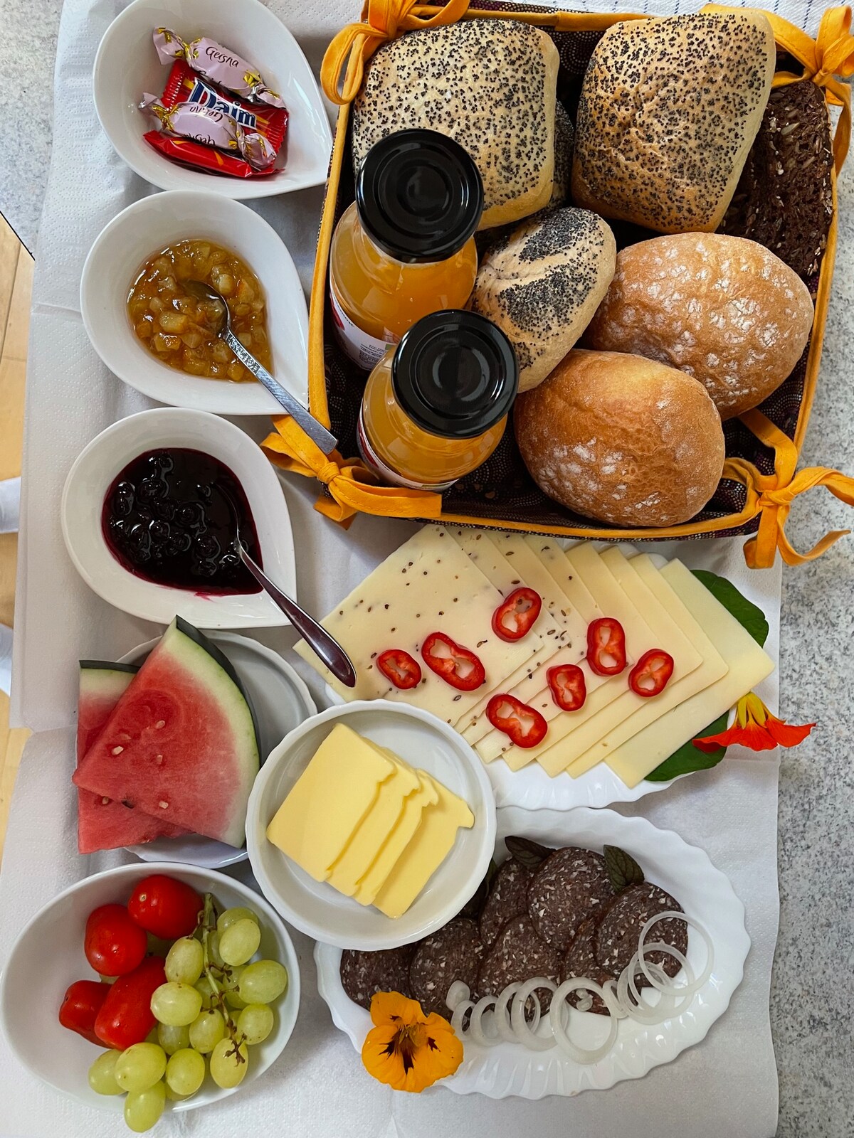 An inviting breakfast tray is arranged with an assortment of items. Fresh rolls with sesame seeds and plain varieties are included, alongside cheeses, spreads, and jams. A selection of fruits, including watermelon, grapes, and cherry tomatoes, is presented, complemented by drinks in glass bottles.