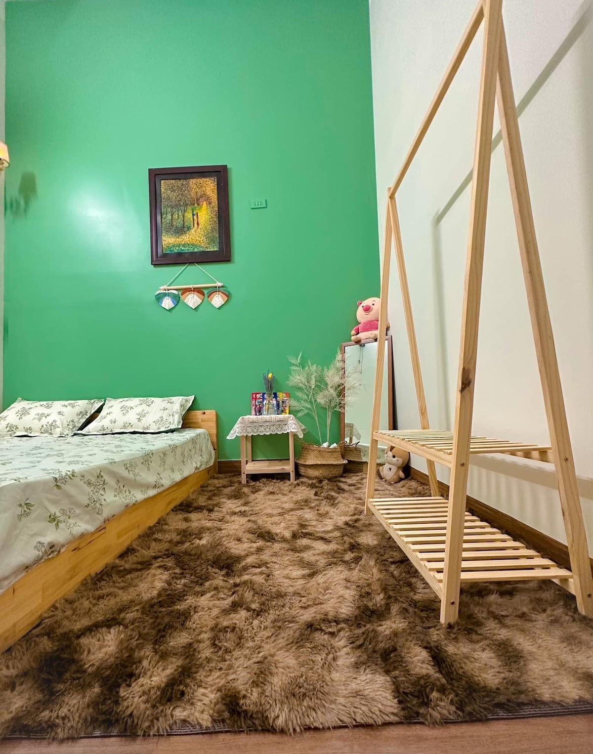 A cozy bedroom features a spacious bed with floral bedding against a vibrant green wall. A wooden shelf stands near the bed, holding decorative items and a plush teddy bear. A soft, brown rug covers the floor, adding warmth to the space.