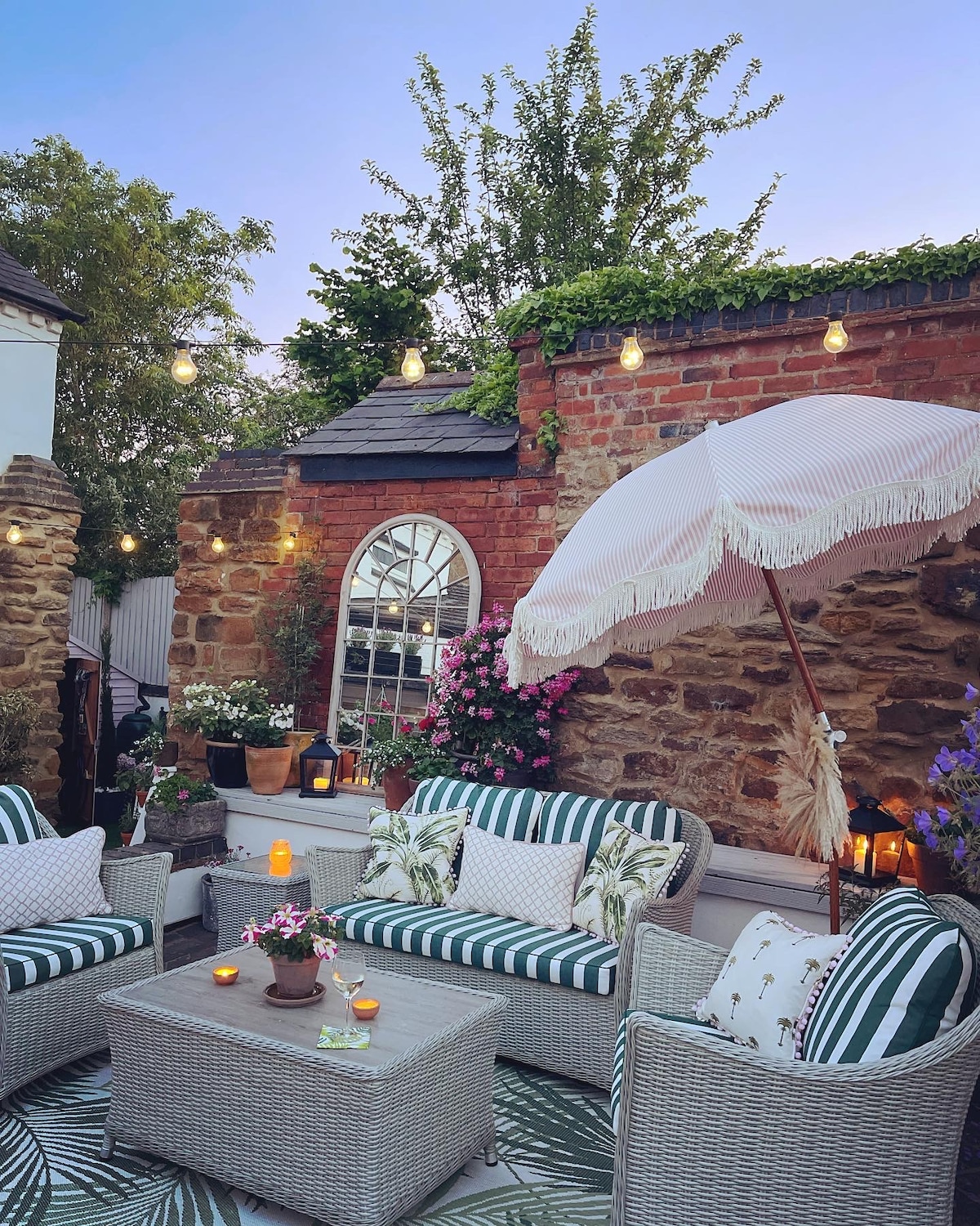 An outdoor seating area is highlighted by woven furniture adorned with green and white striped cushions. A round table sits in the center, surrounded by flowers in terracotta pots. Soft lighting enhances the space, complemented by a large umbrella providing shade.