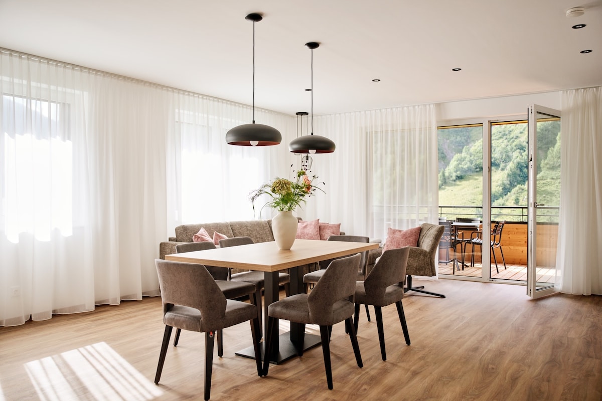 The living area features a wooden dining table surrounded by upholstered chairs, with a cozy sofa positioned nearby. Large windows adorned with light curtains allow natural light to fill the space, while an open door leads to a terrace showcasing mountain views.