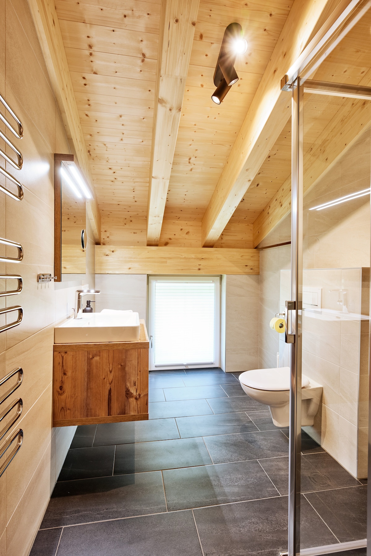 The bathroom features a modern design with wooden accents and a sloped ceiling. A glass shower enclosure is paired with a vanity that includes a sink and storage. A window allows natural light, while a heated towel rack provides comfort.