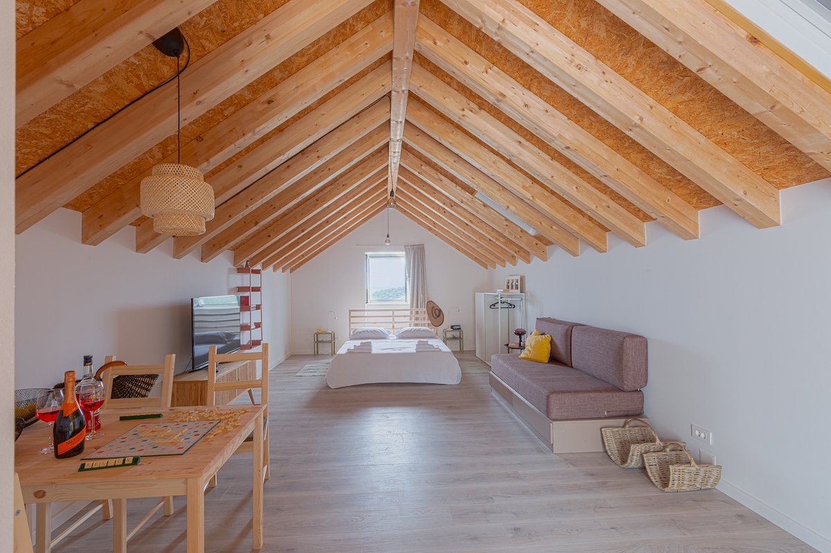 A spacious interior features high, angled wooden beams overhead. A cozy sleeping area includes a large bed, complemented by a sofa and dining table. Natural light illuminates the room from a window with a soft curtain, enhancing the open and airy feel.